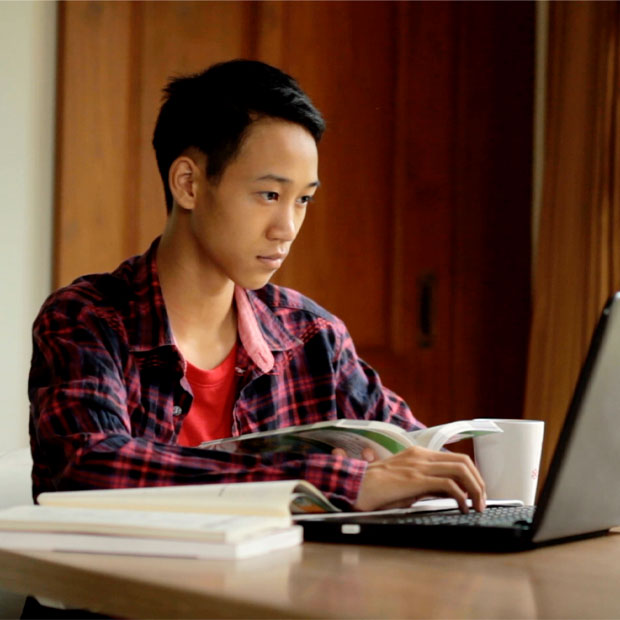 Male student using a laptop while holding a book